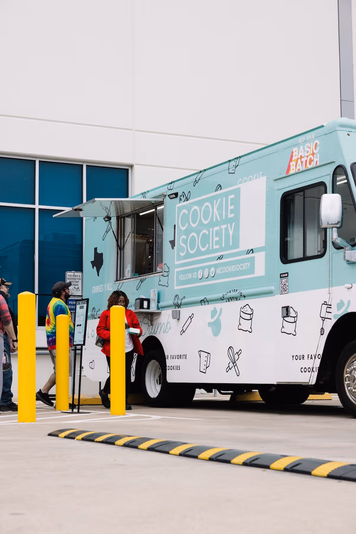 People line up at a blue and white Cookie Society food truck outside a large building.
