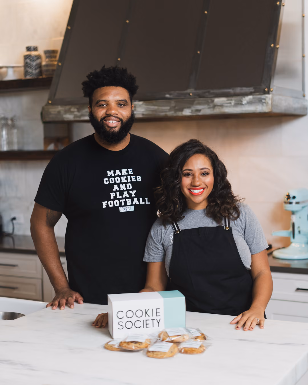 Two people smiling at a kitchen counter with a Cookie Society box and cookies