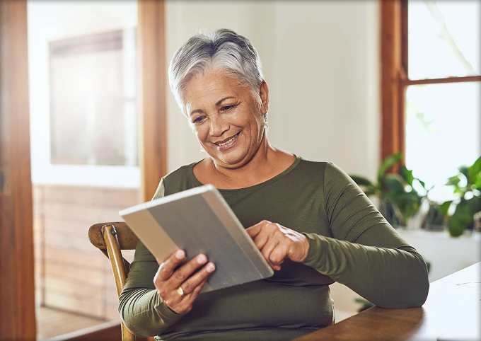 Smiling retired woman sitting at table using a tablet device in a bright room with plants.