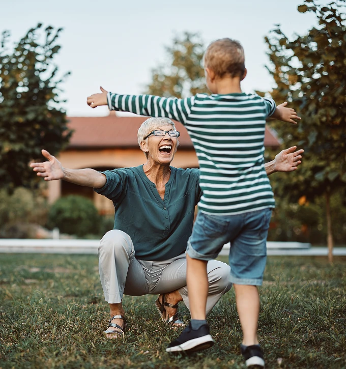 Smiling retired woman crouching outdoors with open arms welcoming a running child in a striped shirt.
