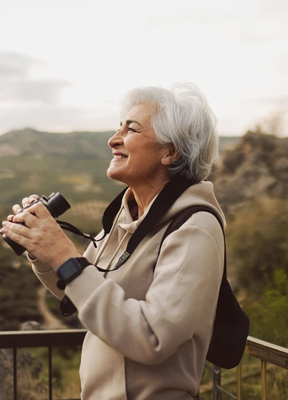 Smiling retired woman holding binoculars outdoors with scenic hills in background.
