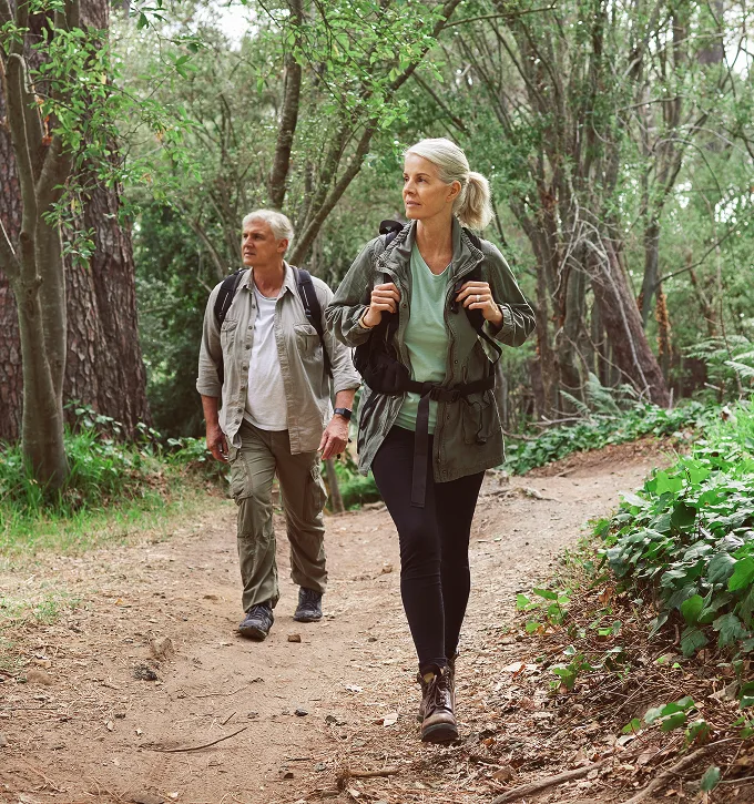 Retired couple hiking on a forest trail surrounded by trees and greenery.