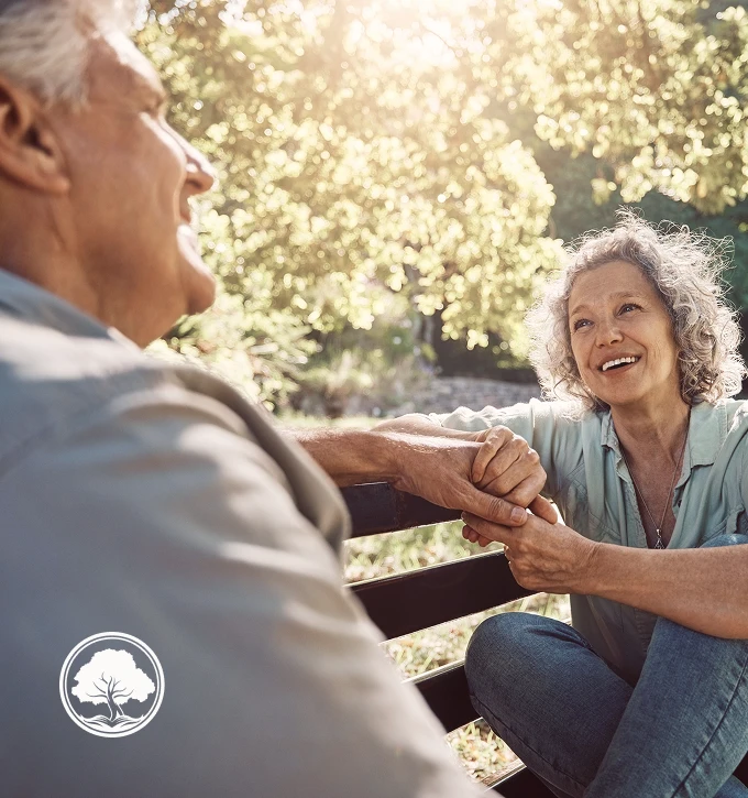 Smiling retired woman holding hands with an retired man on a park bench in sunlight.