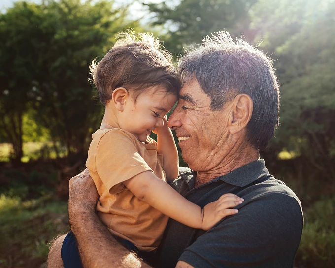 Smiling retired man holding a young child forehead to forehead outdoors with sunlight and trees.