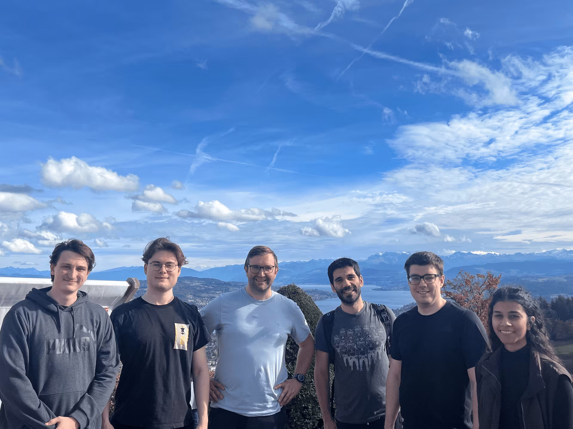 Six people standing outdoors with mountains and lake in the background under a partly cloudy sky.
