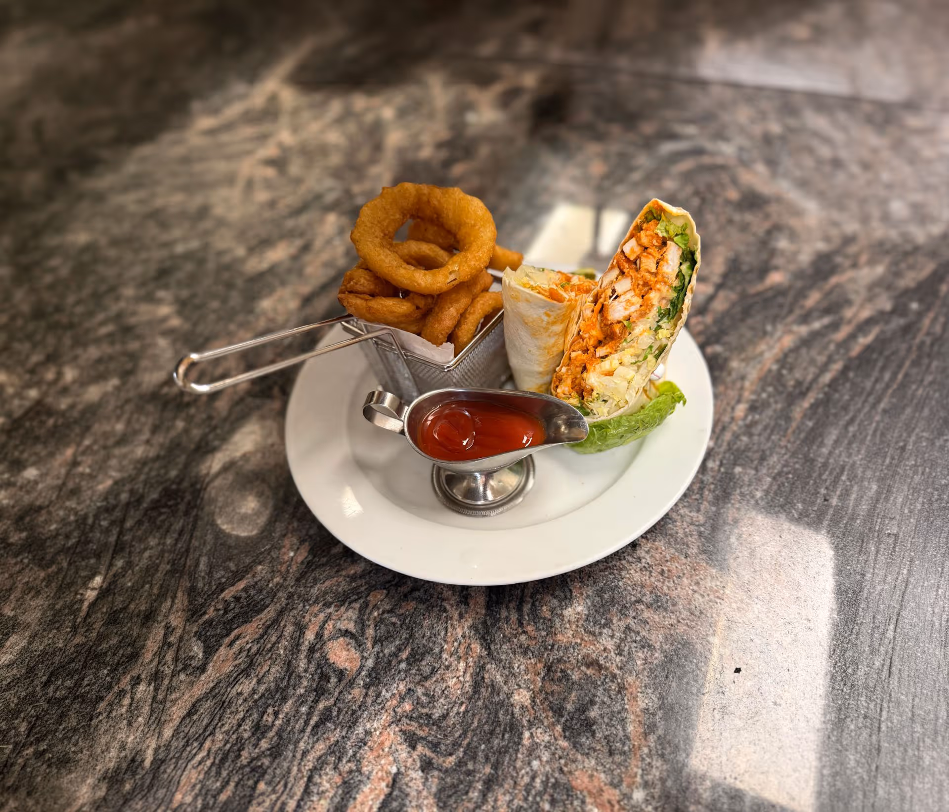 Plate with two chicken wraps, crispy onion rings in a metal basket, and a sauce cup of ketchup.