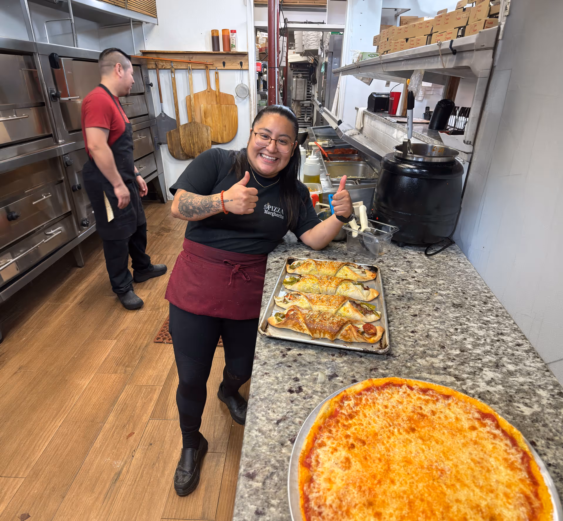 Smiling woman giving thumbs up by counter with pizza and calzones in pizzeria kitchen.