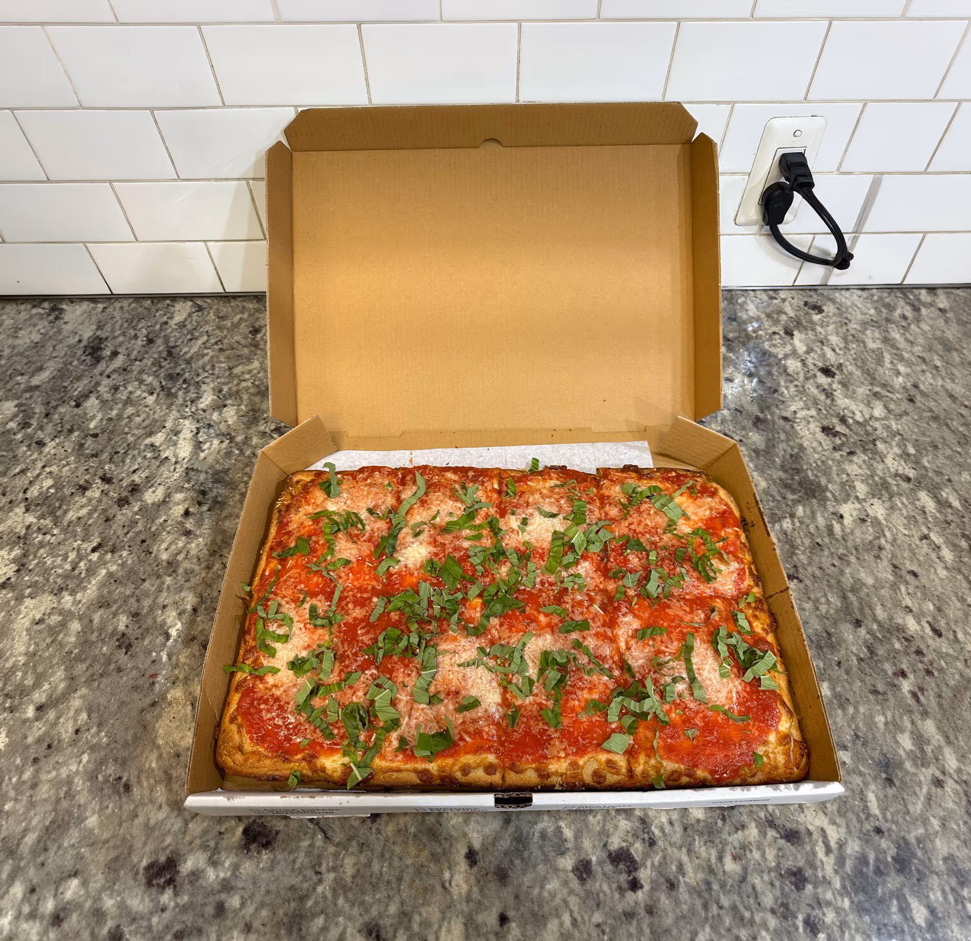 Rectangular pizza with tomato sauce, cheese, and fresh basil leaves in an open cardboard box.