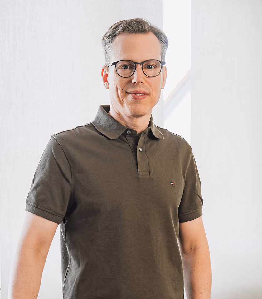 Mann mit Brille und olivgrünem Polohemd vor hellem Hintergrund.