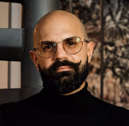Man with a bald head, full beard and curled mustache wearing round glasses and a black turtleneck.