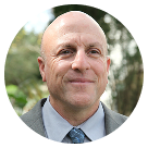 Smiling bald man wearing a suit and tie outdoors with blurred trees in the background.