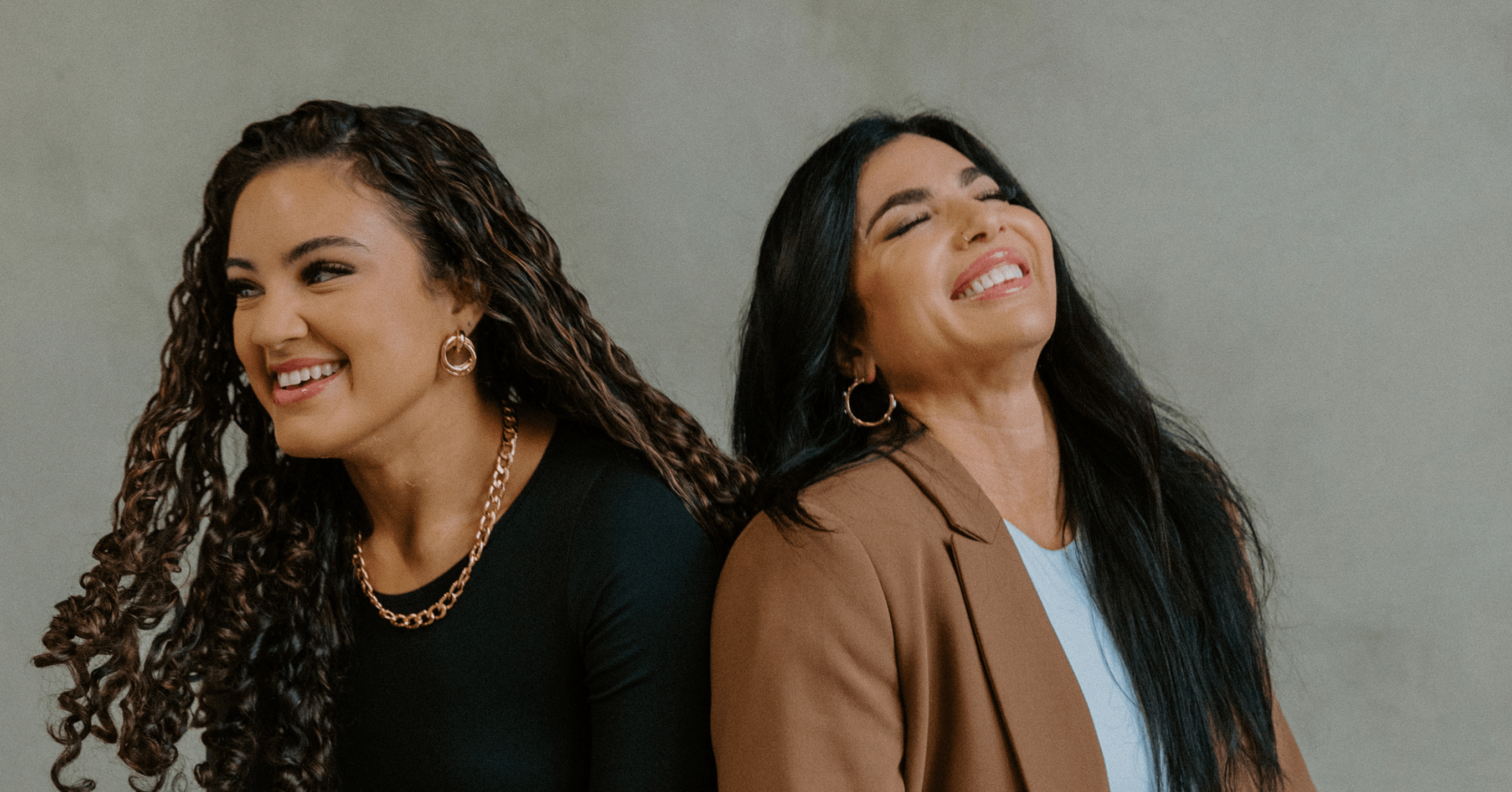 Two smiling women with long dark hair, one in a black top and one in a brown blazer, against gray background.