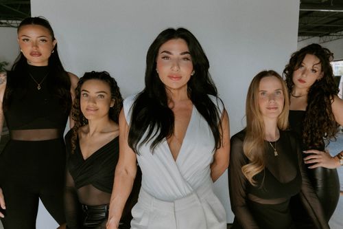 Five diverse women dressed in stylish black outfits walking together indoors against a gray wall with green plants on each side.