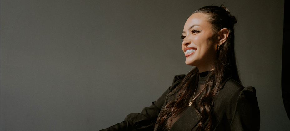 A facial specialist woman with long dark hair wearing a black outfit smiling against a dark gray background.