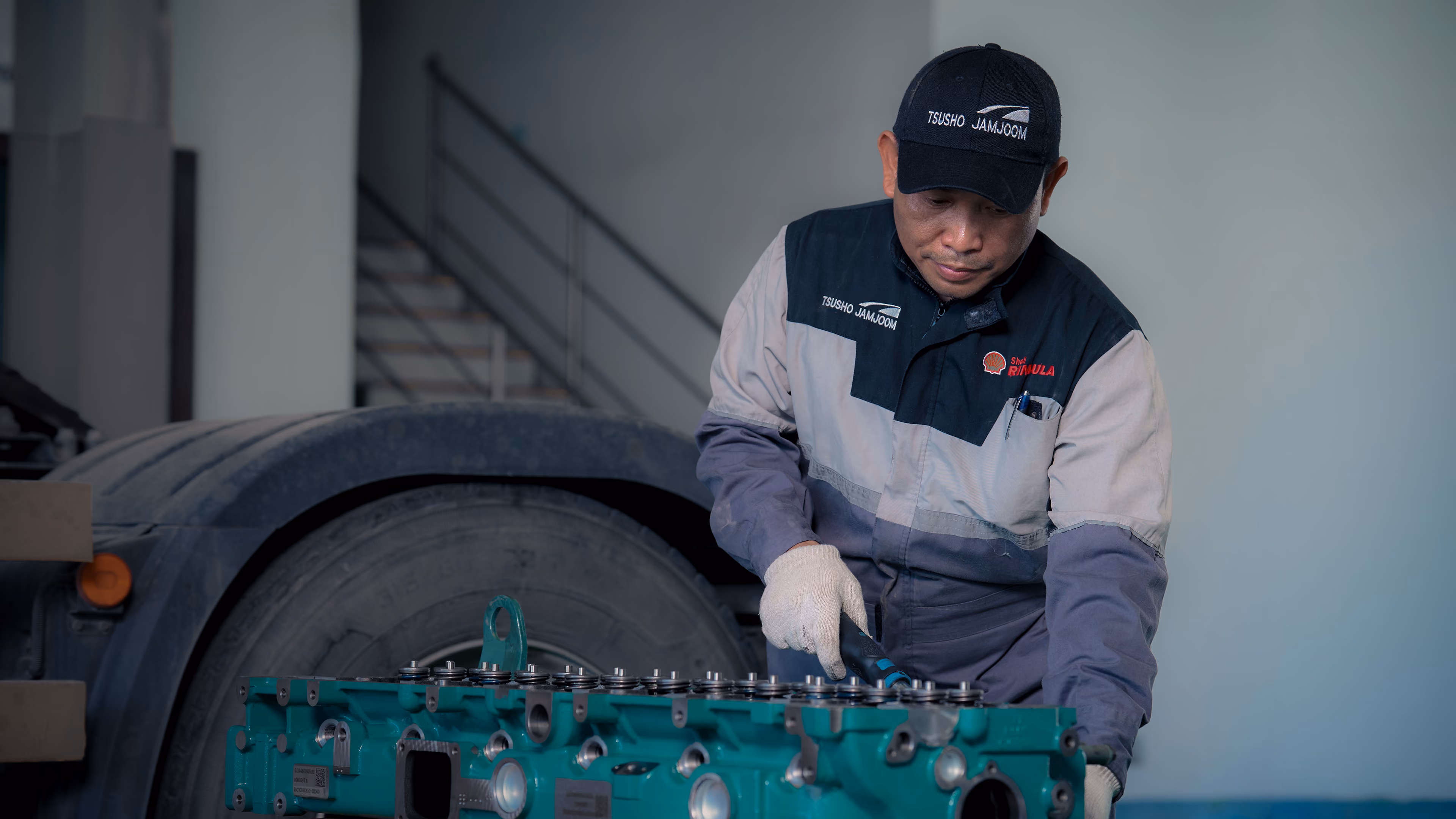 Mechanic in blue uniform holding tablet in a truck service garage with a white truck on the lift.