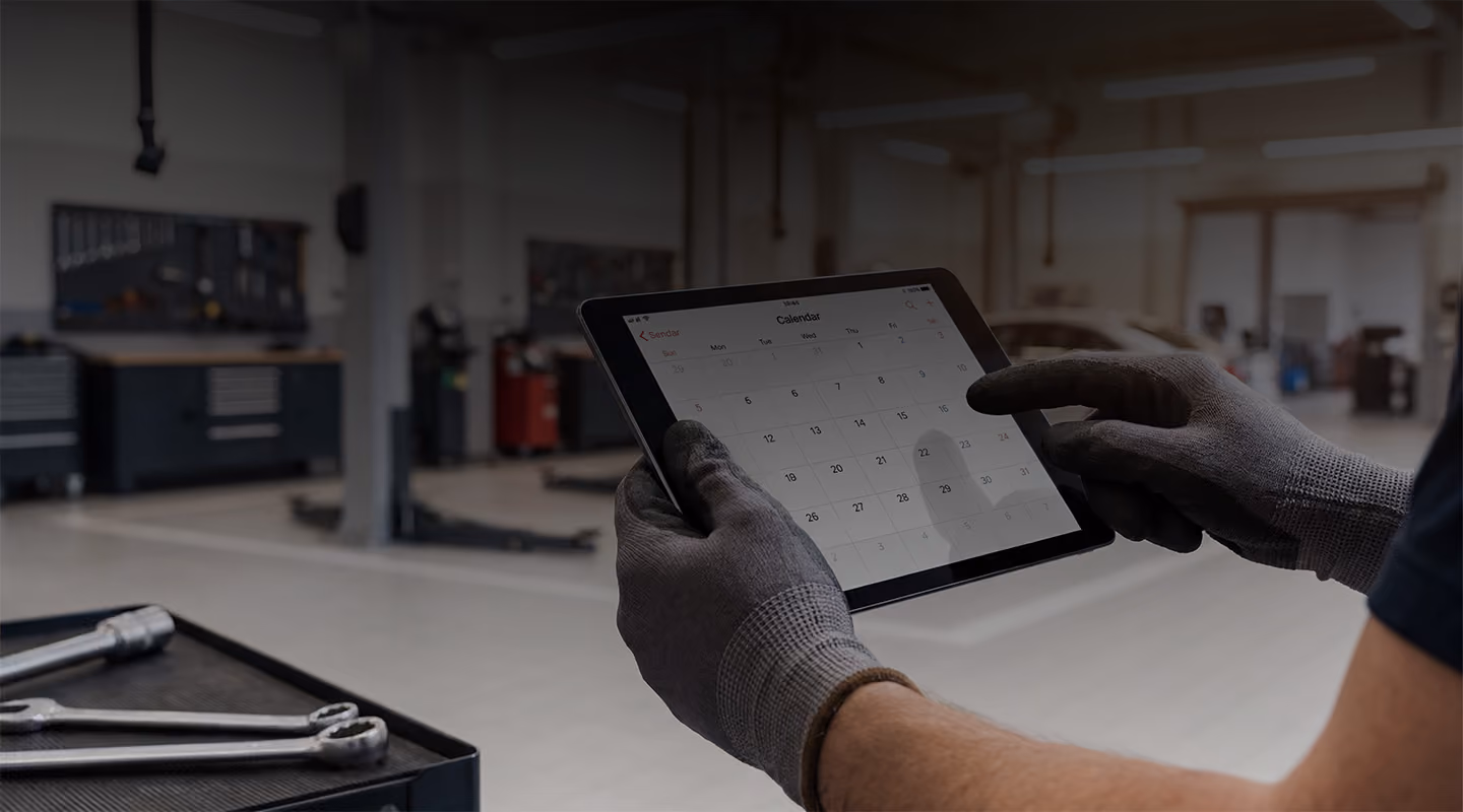 Person wearing work gloves using a tablet displaying a calendar in a garage or workshop.