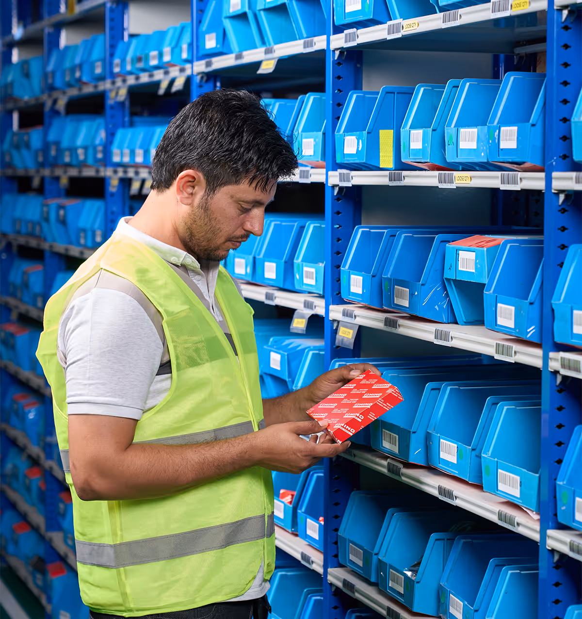 Man in reflective vest inspecting a red box among blue storage bins on metal shelves in a warehouse.