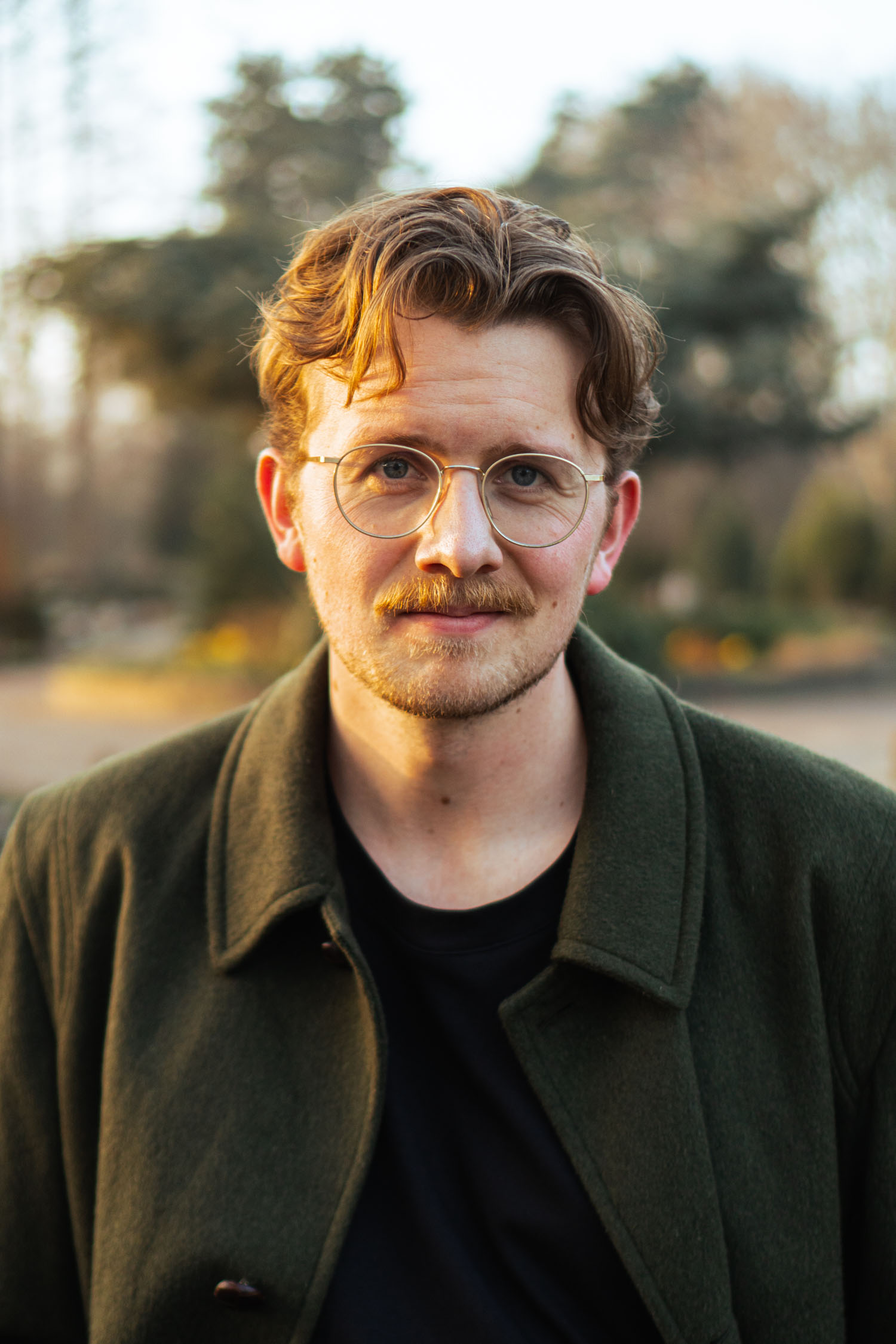 Man with light brown hair, mustache, round glasses, wearing green coat and black shirt outdoors.