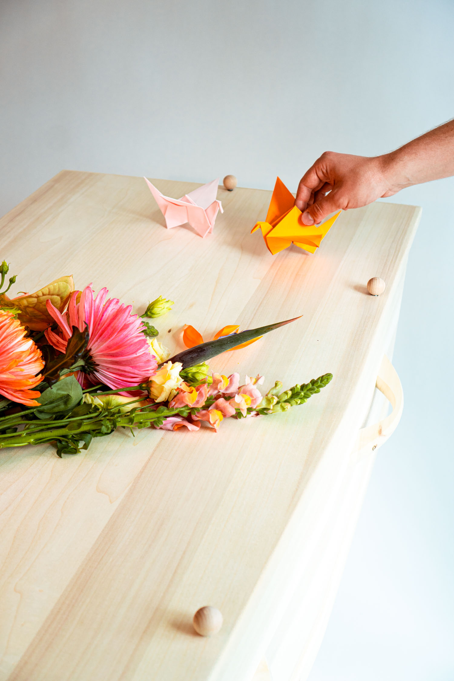 Hand placing orange origami crane beside a pink one on a light wood surface with colorful flowers.