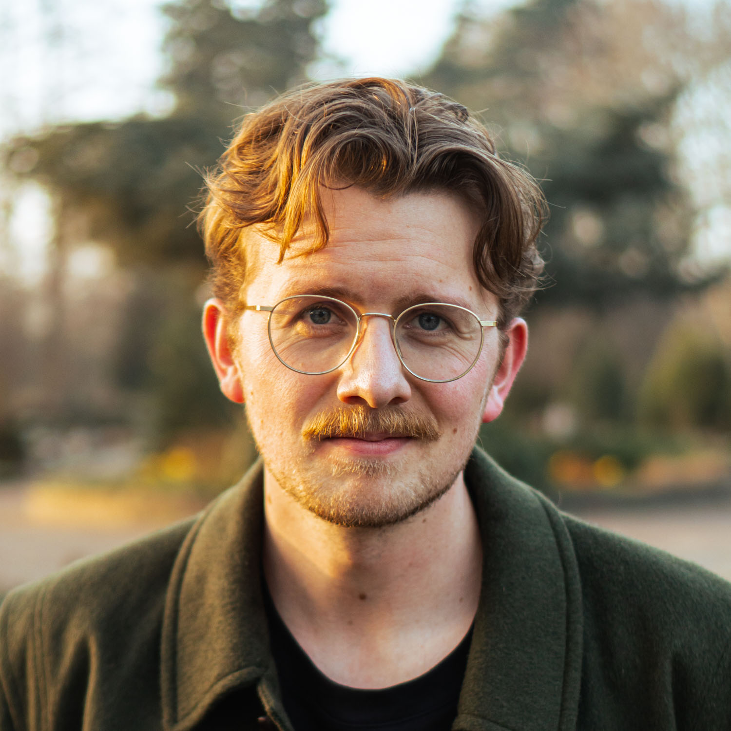 Young man with wavy hair, mustache, round glasses, and green jacket outdoors in soft light.