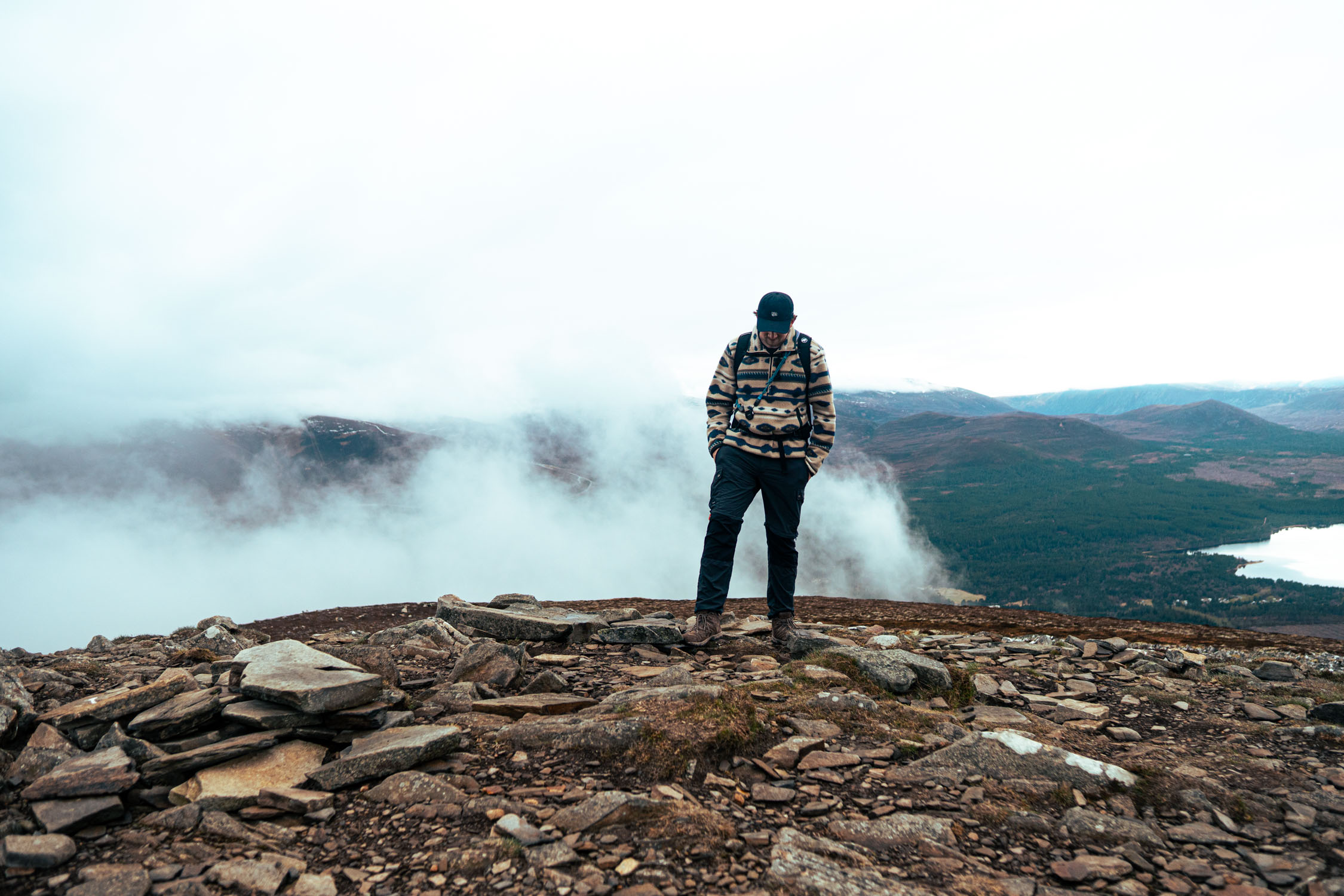 Man in patterned sweater and cap standing on rocky hill with misty mountains and lake in background.