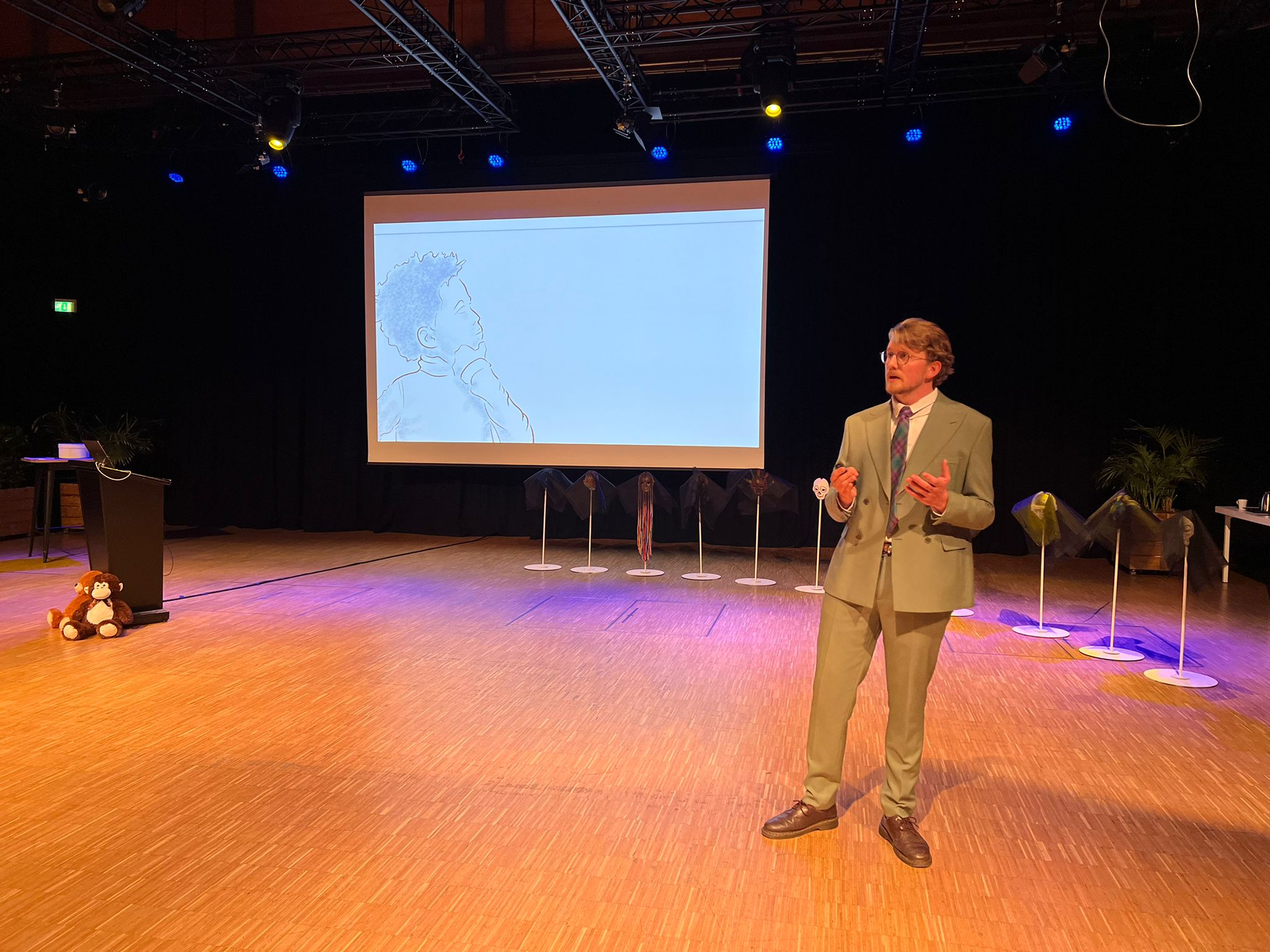 Man in pak geeft presentatie op podium met projectie van kindertekening achter hem.