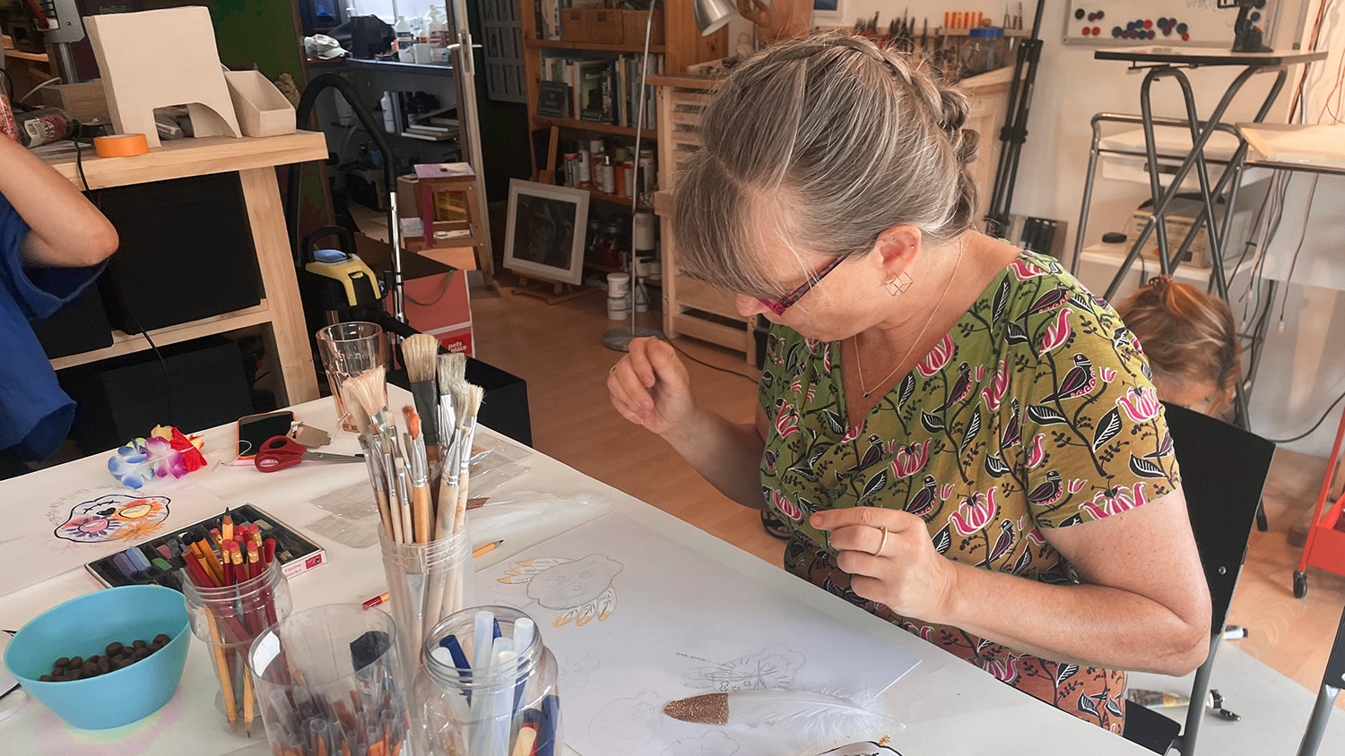 Vrouw tekent creatief aan tafel met potloden, penselen en kunstbenodigdheden in atelier.