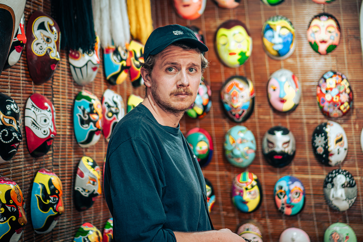 Man met zwart shirt en pet staat in een winkel vol kleurrijke, traditionele maskers aan de muur.