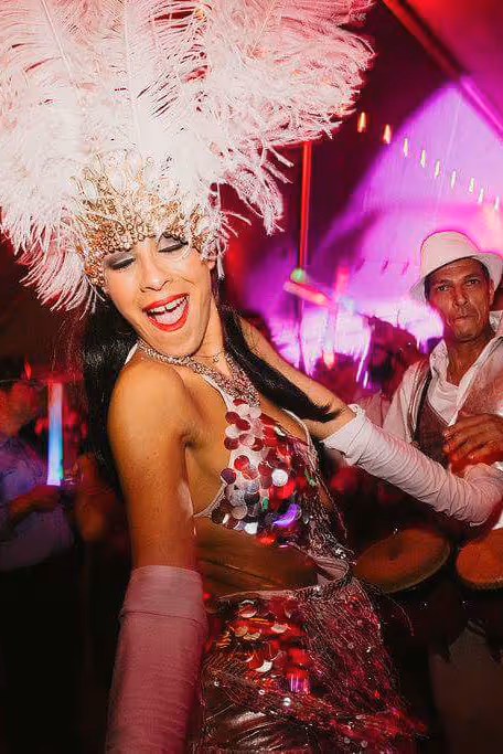 Woman dancing joyfully at a party, wearing a silver sequin dress and large white feathered headpiece.