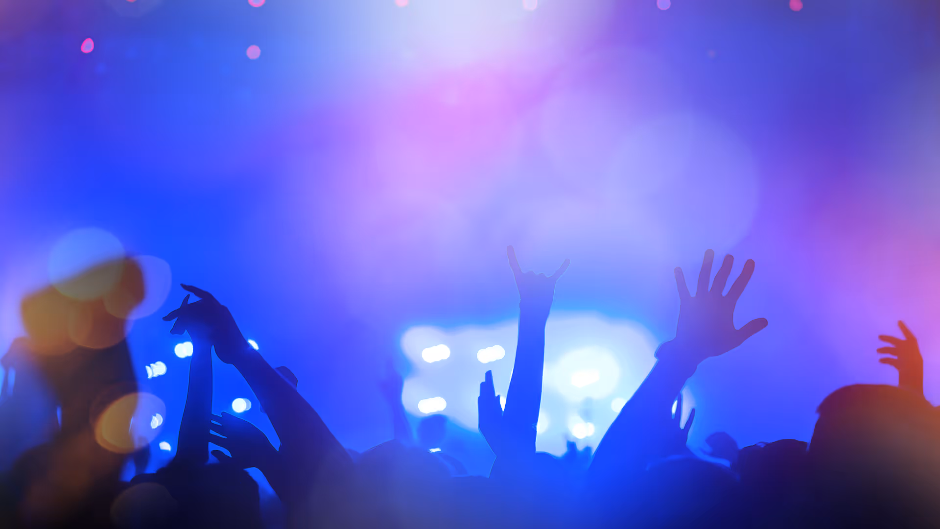 Silhouettes of raised hands at a concert with blue and purple stage lights in the background.