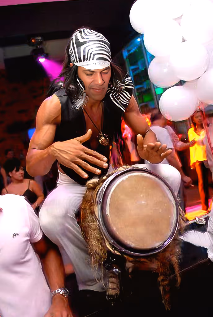Man wearing a black vest and striped headscarf playing a drum at a lively party.