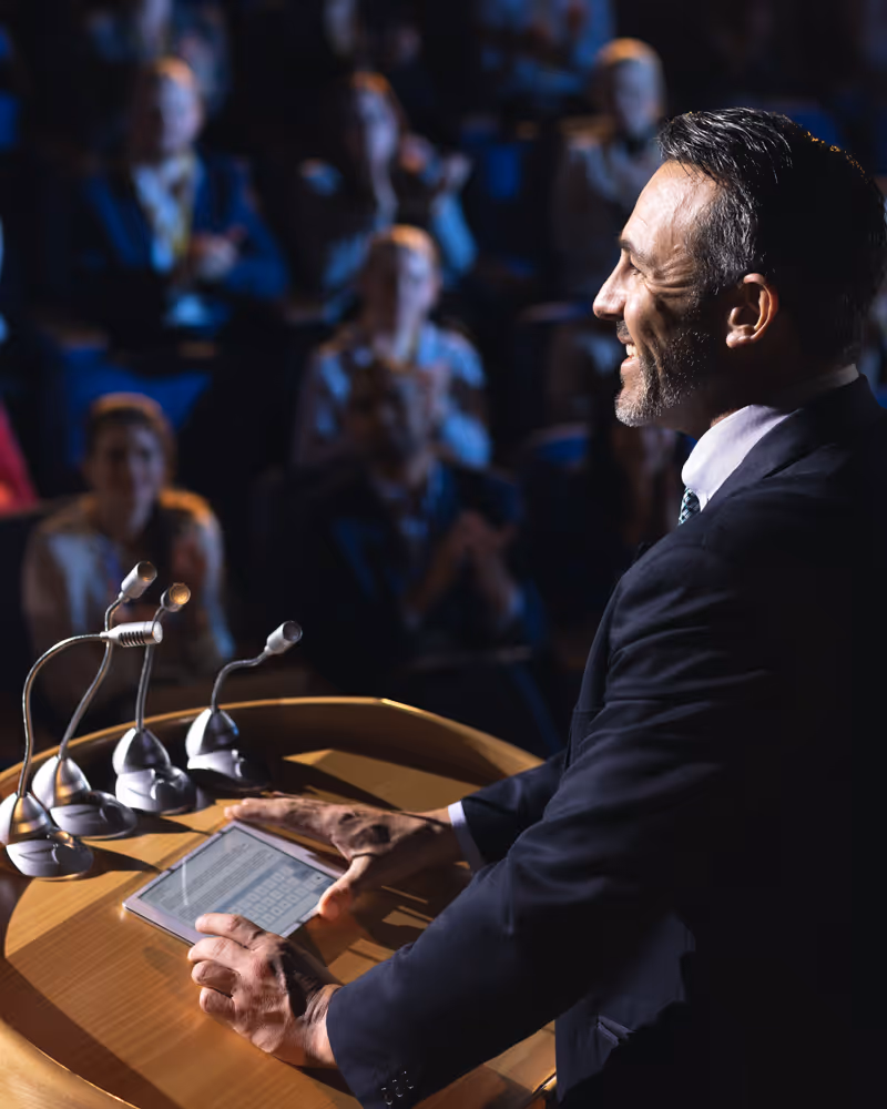 Smiling man in suit speaking at podium with microphones and tablet, audience clapping in background.