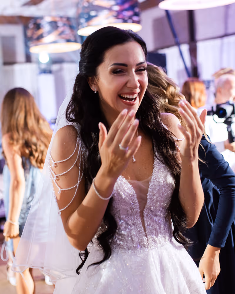 Smiling bride in a sparkling wedding dress laughing and clapping her hands at a wedding reception.