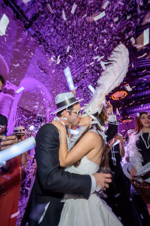 Couple in festive hats kissing amid confetti at a lively party with purple lighting.