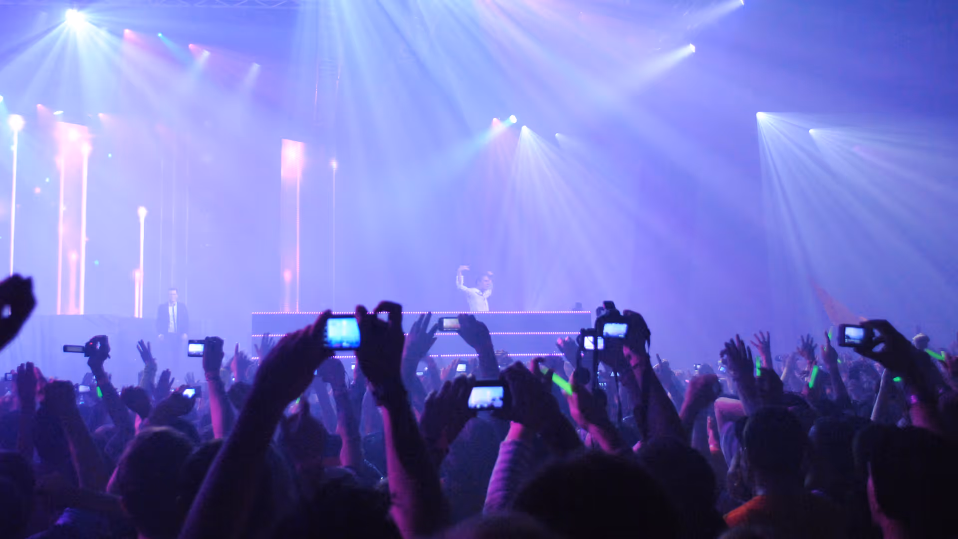Crowd at a concert holding phones up to record the DJ on stage under colorful lighting.