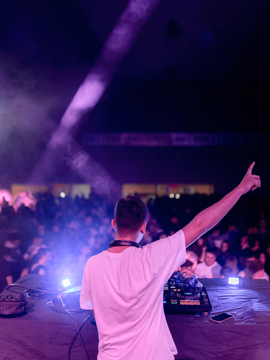 DJ wearing headphones raising hand while playing music for a crowded, purple-lit audience.