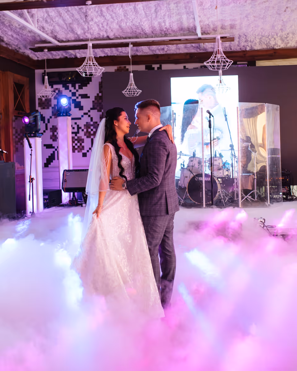 Bride and groom dancing on a fog-covered dance floor at their wedding reception with stage lights behind.