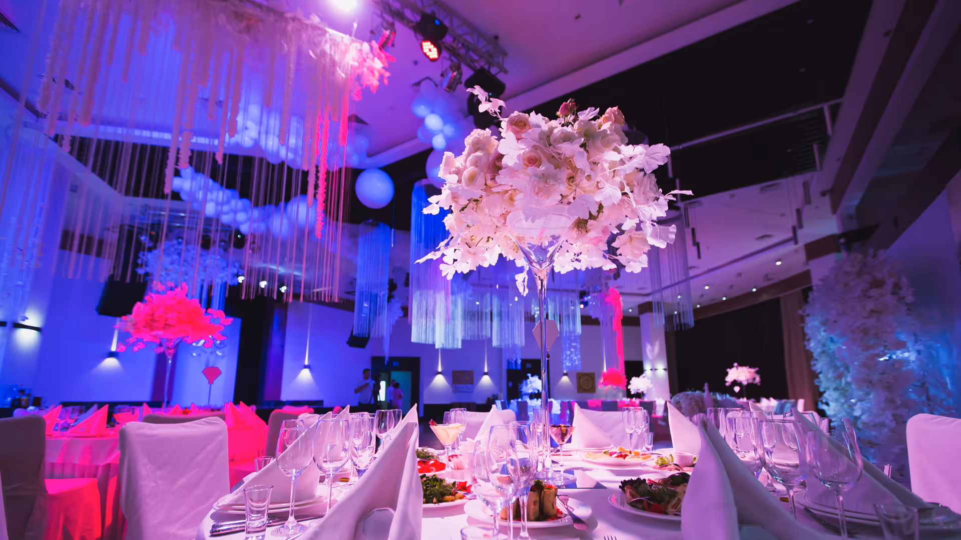 Elegant wedding ballroom with tables set and tall floral centerpieces under purple and pink lighting.