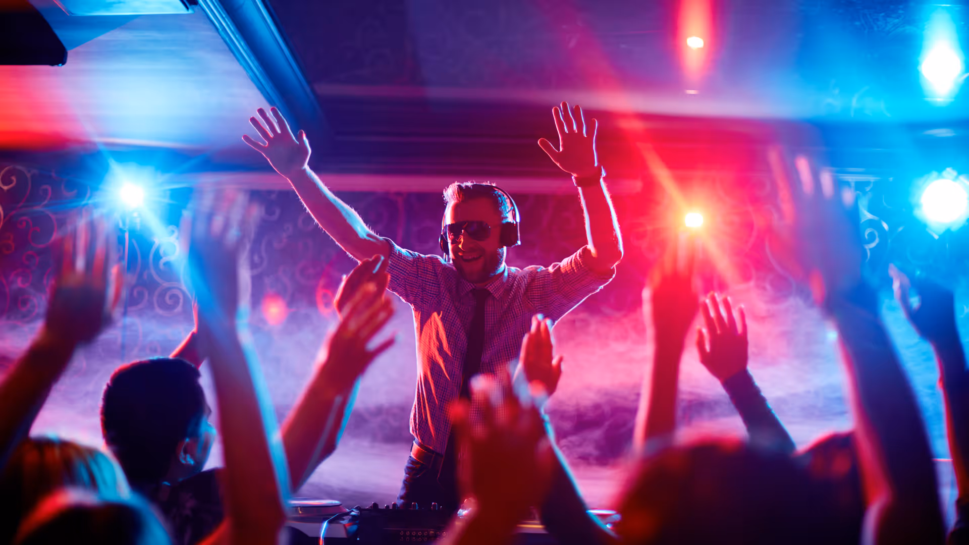 DJ wearing headphones and sunglasses with hands raised, crowd reaching up under colorful lights.