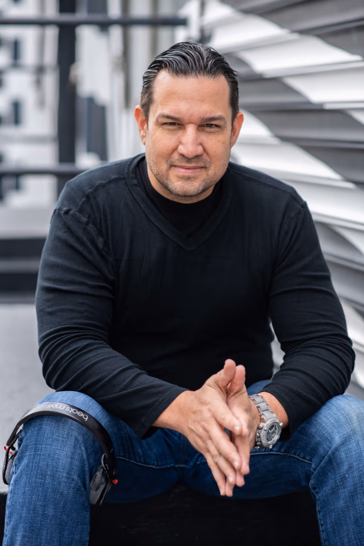 Man with slicked-back hair wearing a black shirt and blue jeans sitting with clasped hands.