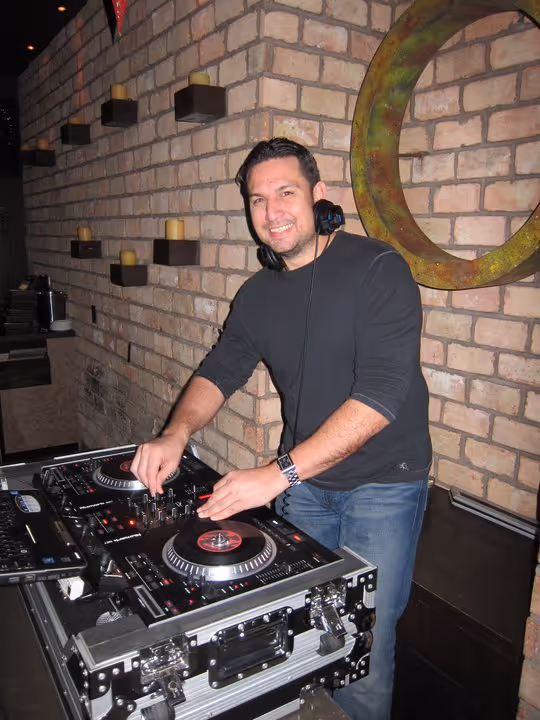 Smiling DJ wearing headphones mixes music on turntables at a brick wall with candles.
