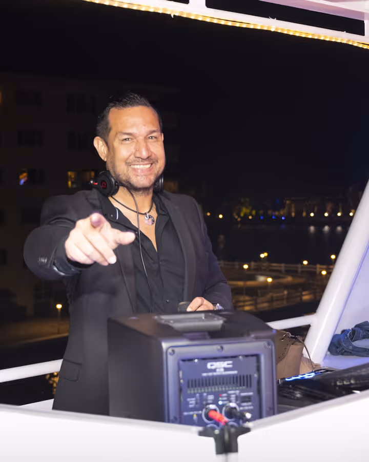 Smiling DJ wearing headphones points forward while standing behind DJ equipment at night outdoors.