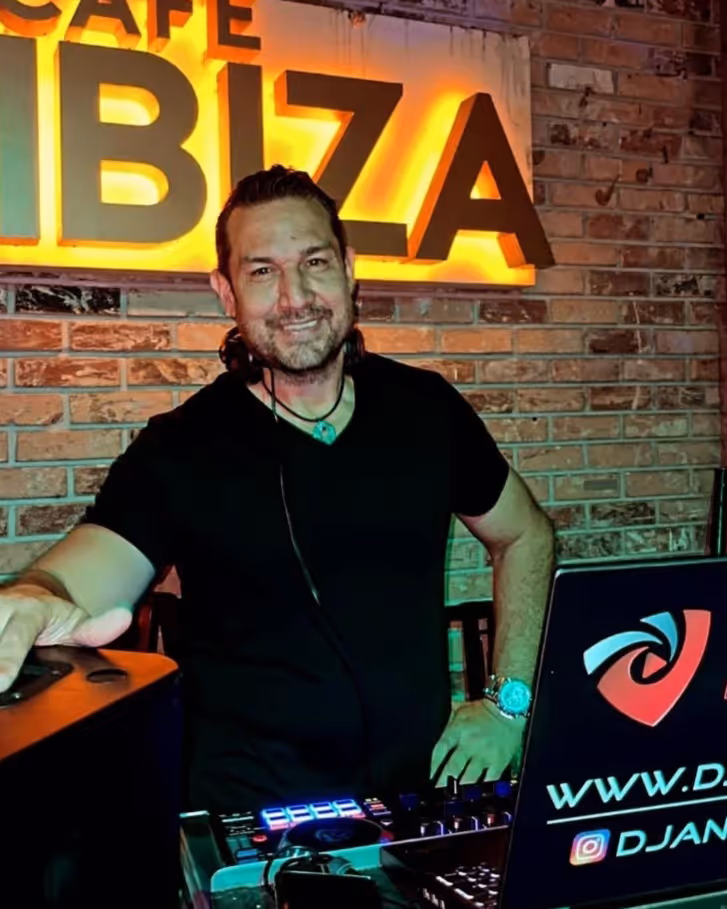 Smiling DJ with headphones stands behind mixing console and laptop at Cafe Ibiza with brick wall.