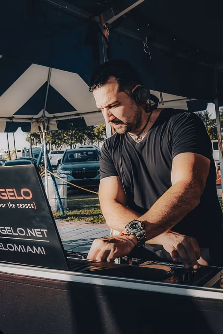 Male DJ wearing headphones using a mixer under a tent with cars parked in the background.
