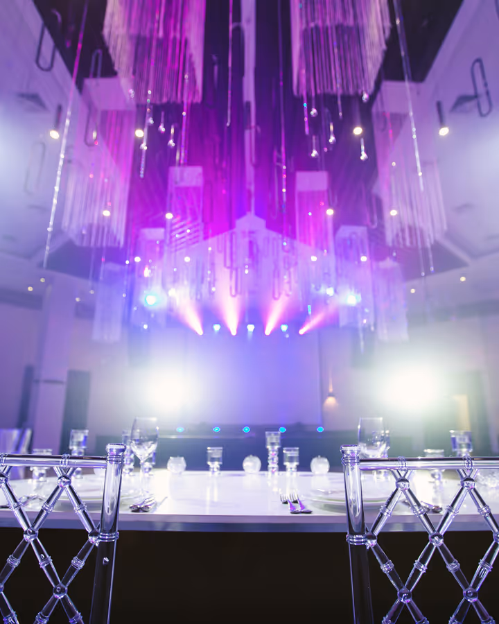 Elegant dining setup with clear chairs and stage lighting casting purple hues in event venue.