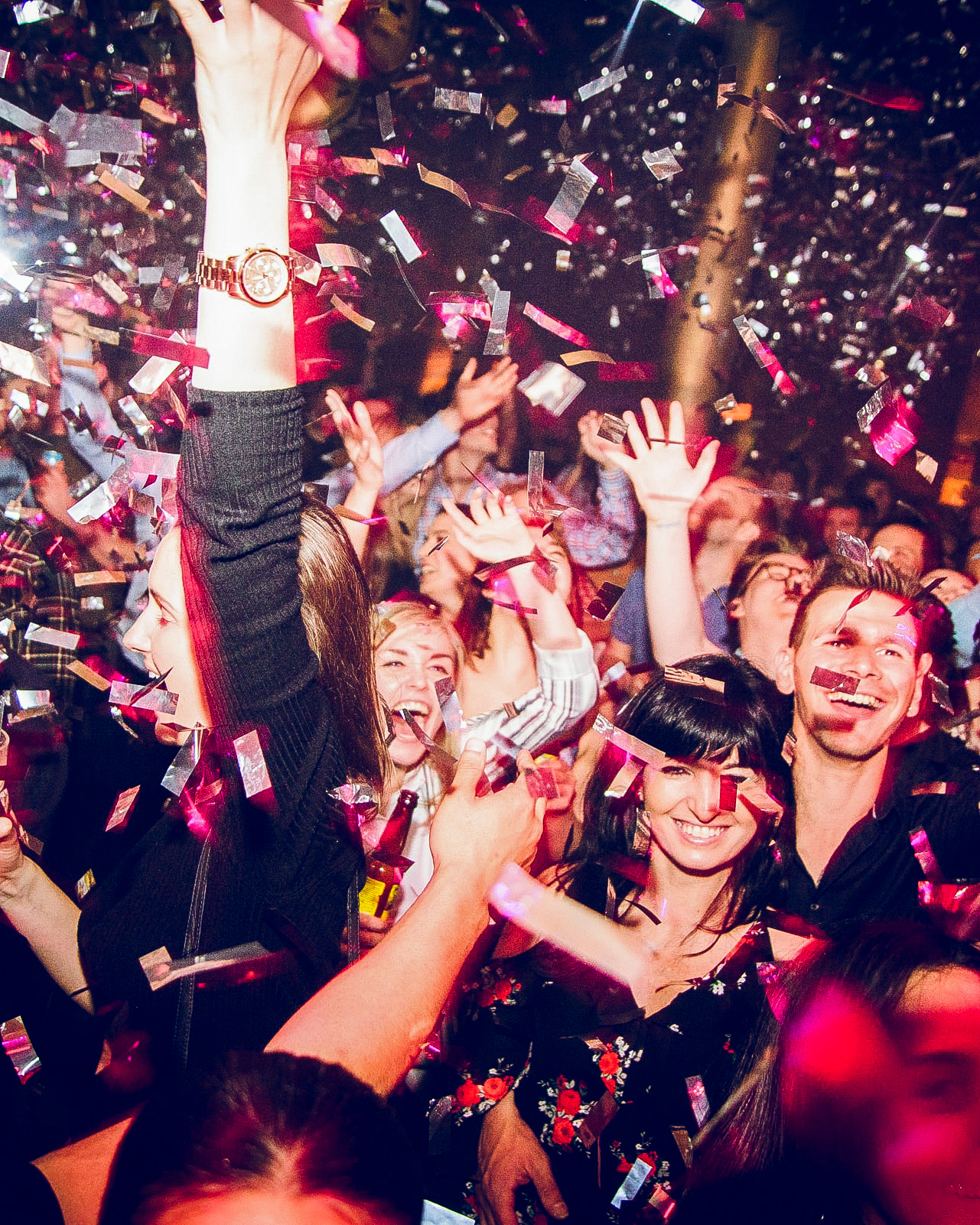 Crowd at a party celebrating with confetti falling and people raising their hands, smiling and cheering.