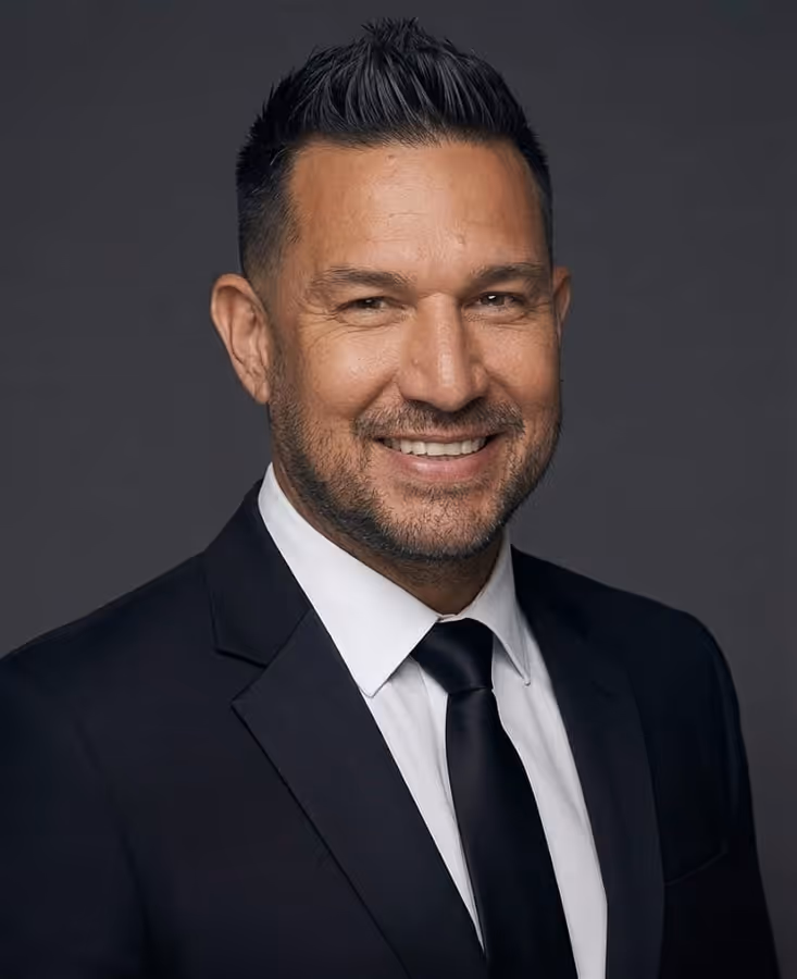 Dj Angelo smiling with short dark hair wearing a black suit, white shirt, and black tie against a gray background.