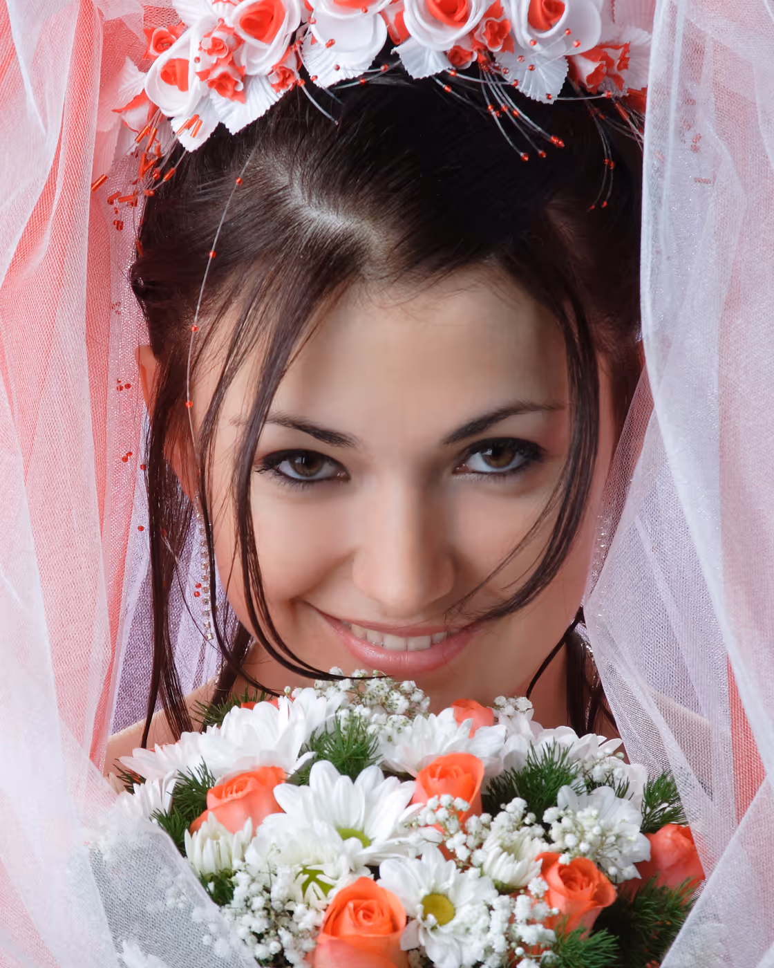 Smiling bride with dark hair wearing a floral crown and veil holding a bouquet of white and coral flowers.