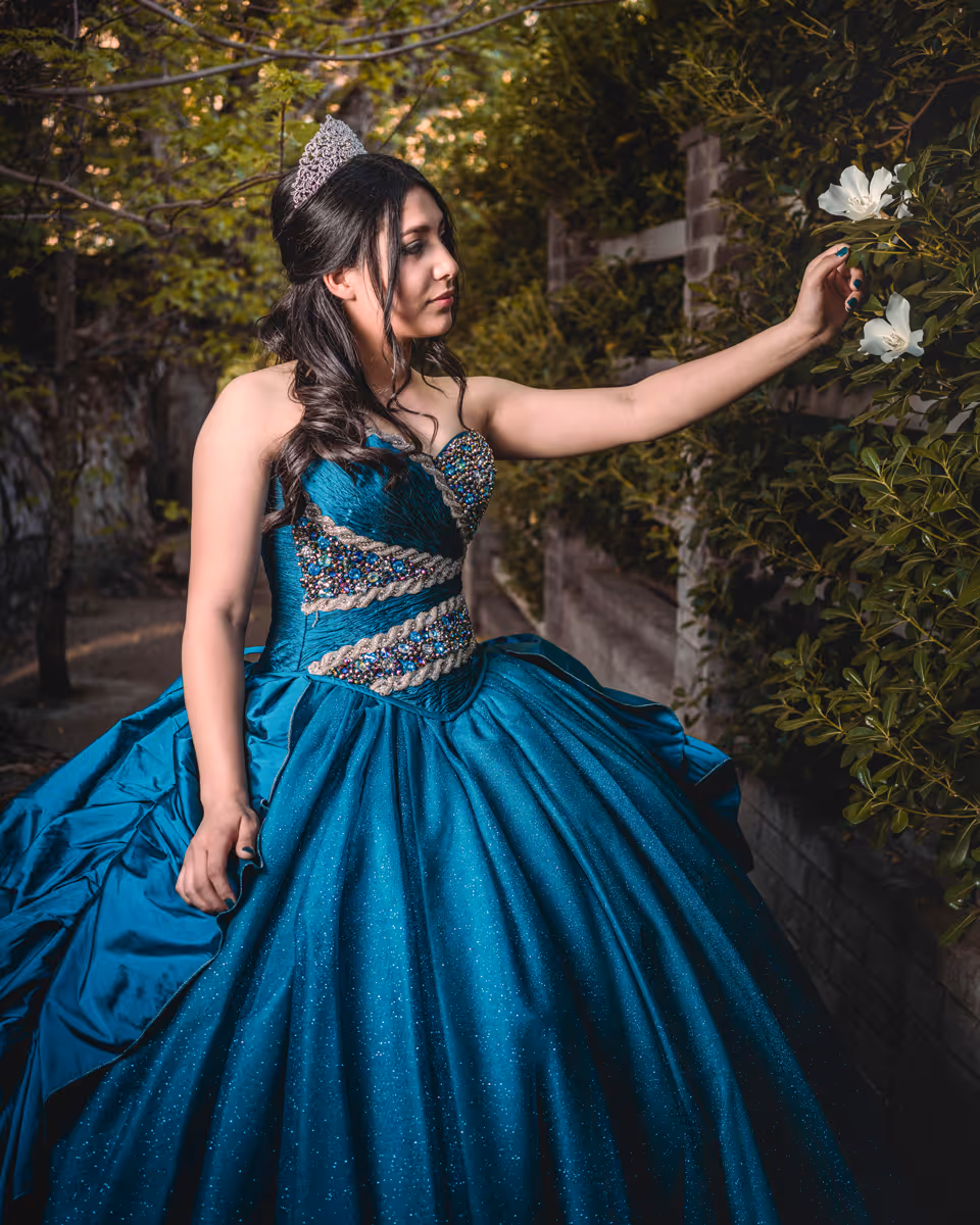 Young woman in blue embellished quinceañera dress and tiara touching white flowers on a bush.