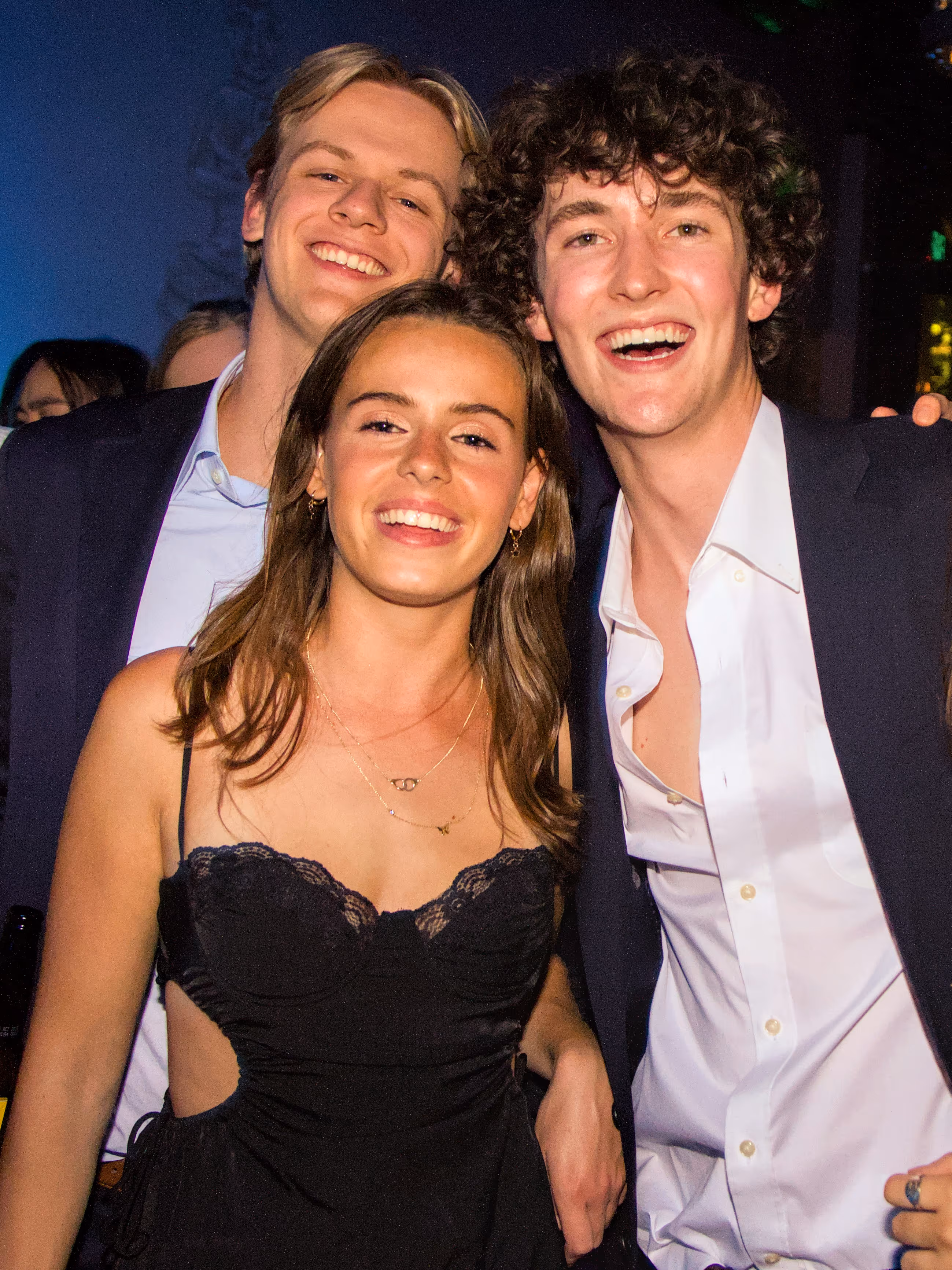 Three young adults smiling and posing closely together at a social event with dark background.